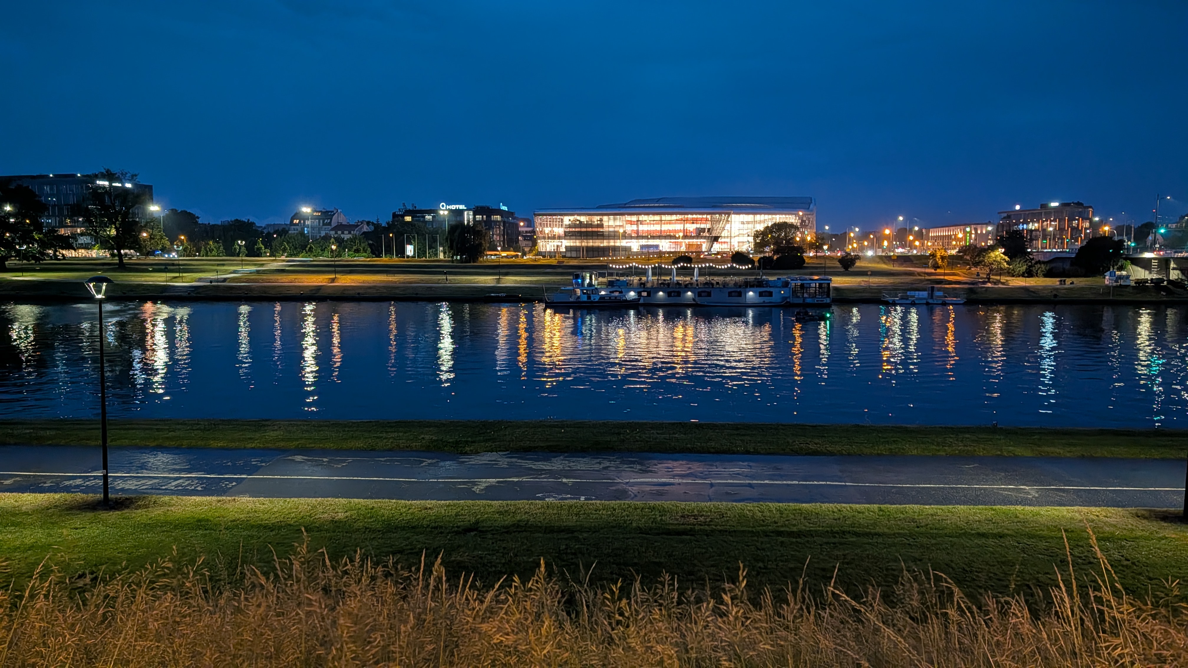 Nighttime view across a river with city lights reflecting on the water, a boat docked near the shore, and a brightly lit modern building in the background.