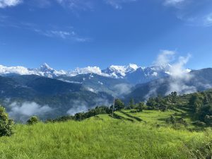 Snow-capped mountains under a clear blue sky with scattered clouds. In the foreground, lush green hills with patches of trees convey a serene landscape.