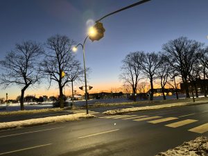 A wide shot of a city street intersection at dusk under a deep blue sky transitioning to a bright orange sunset. An asphalt road with white crosswalk stripes is flanked by snow-covered ground and a line of tall, leafless winter trees. A yellow traffic light and street lamp glow brightly in the foreground. 