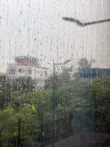 Raindrops trail down a glass pane, blurring the view of a green landscape and distant buildings. 