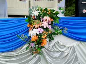 A decorative floral arrangement of artificial orange, pink, white, and lavender chrysanthemum-style blooms with green foliage, mounted at the center of a draped blue and white satin fabric table skirt at an outdoor event, Nepal.