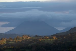 A distant volcano rises behind green hills and forest, partially hidden by thick blue-gray clouds. Warm sunlight highlights parts of the foreground, creating contrast with the dark, misty mountain in the background.