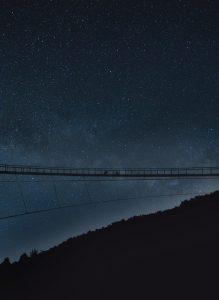 A silhouetted suspension bridge spans across a star-filled night sky. The Milky Way is visible, creating a serene and awe-inspiring atmosphere.