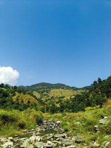 Clear blue sky over a lush, green mountain landscape. A rocky stream flows in the foreground, adding a serene, natural vibe to the scene.