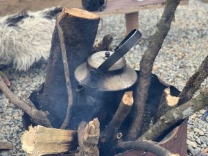 Metal kettle placed among firewood, smoke rising from smoldering wood.