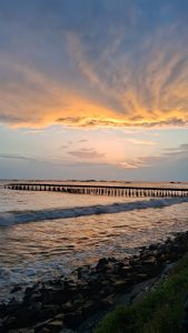 ​A scenic sunset view over the ocean with a long wooden pier stretching across the water, gentle waves breaking on a rocky shoreline in the foreground, and golden-orange clouds filling the sky.
