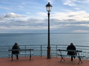 Two people sitting on their backs on two different benches facing the sea, separated by a street lamp