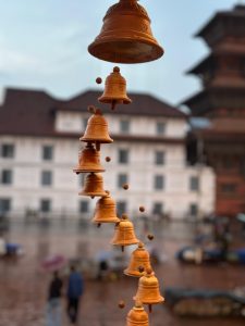 Close-up of terracotta bells hanging in a line, with a blurred background of historical architecture and people walking on a wet day. The scene feels serene and traditional.