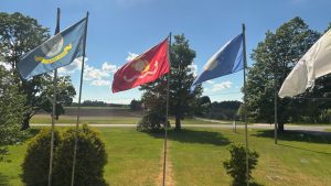 A row of government flags on a lawn seen from inside a building. In the distance is a plowed field.