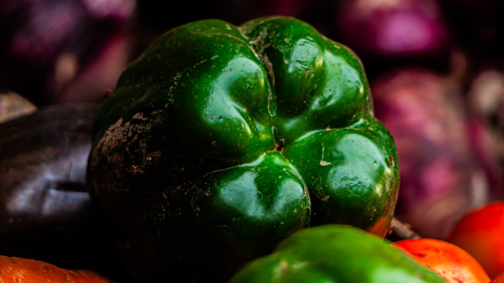 Close-up macro shot of a large green bell pepper, showcasing its glossy, lobed texture with red tomatoes and purple onions softly blurred in the background.