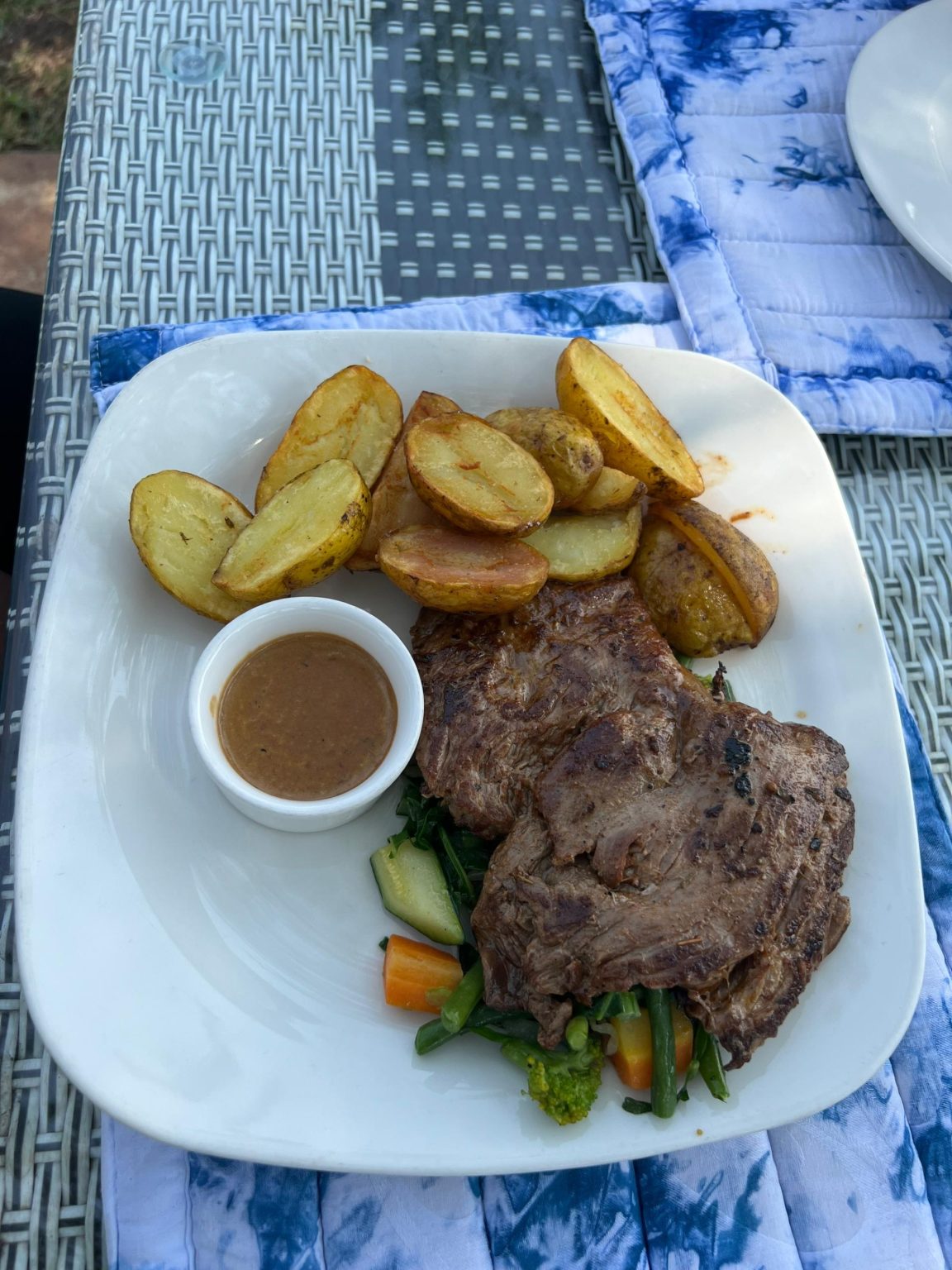 A grilled beef steak platter served with roasted potato wedges, a small white cup of brown sauce, and a bed of sautéed mixed vegetables including carrots, zucchini, and broccoli.