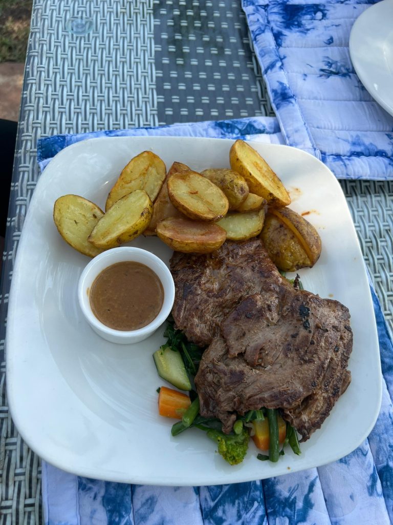 A grilled beef steak platter served with roasted potato wedges, a small white cup of brown sauce, and a bed of sautéed mixed vegetables including carrots, zucchini, and broccoli.