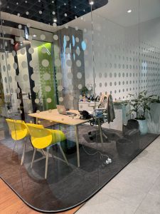Modern glass-walled office with a wooden desk, open laptop, two bright yellow chairs, and potted plants. Minimalist and vibrant ambiance.
