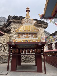 Intricately decorated stupa with colorful carvings and prayer wheels beneath. Surrounded by stone buildings, prayer flags, and mountains in background.
