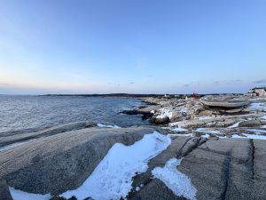 A wide, rocky coastline featuring expansive grey granite formations dusted with patches of white snow. The rugged shore meets a deep blue, undulating ocean under a clear, pale gradient sky that transitions to soft pink on the horizon. A few small coastal buildings are visible in the far distance. 
