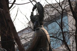 Statue of a person with a feathered hat, featuring a pigeon perched on their shoulder. Bare trees and an urban backdrop create a serene scene.