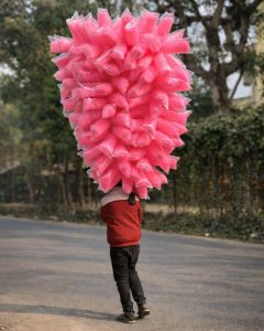 A boy walks along a quiet street lined with trees, carrying a massive bundle of bright pink cotton candy bags. 
