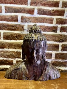 An aged wooden carved Buddha head bust displayed on a wooden surface against a rustic brick wall. 