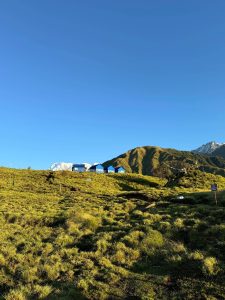 A scenic landscape showing a wide, grassy hill illuminated by sunlight. On the crest of the hill sits a row of small blue structures or tents, set against a backdrop of green mountain ridges and a snow-capped mountain peak poking out on the far right under a vast, bright blue sky. 