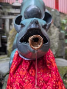 Traditional fox-shaped water fountain at Fushimi Inari Taisha Shrine in Kyoto, with water flowing from a bamboo spout and a red ceremonial cloth. The scene reflects Japanese purification rituals, Shinto traditions, and spiritual devotion.