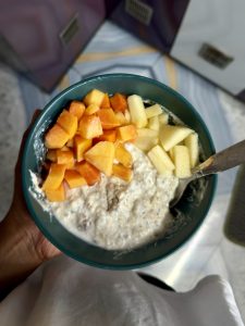 A hand holding a teal bowl of overnight oats topped with diced papaya and sliced apple pieces, Nepal. 