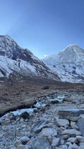 A small stream winds through a rocky mountain valley, with patches of ice along the water’s edge and towering snow-covered peaks rising under a clear blue sky.