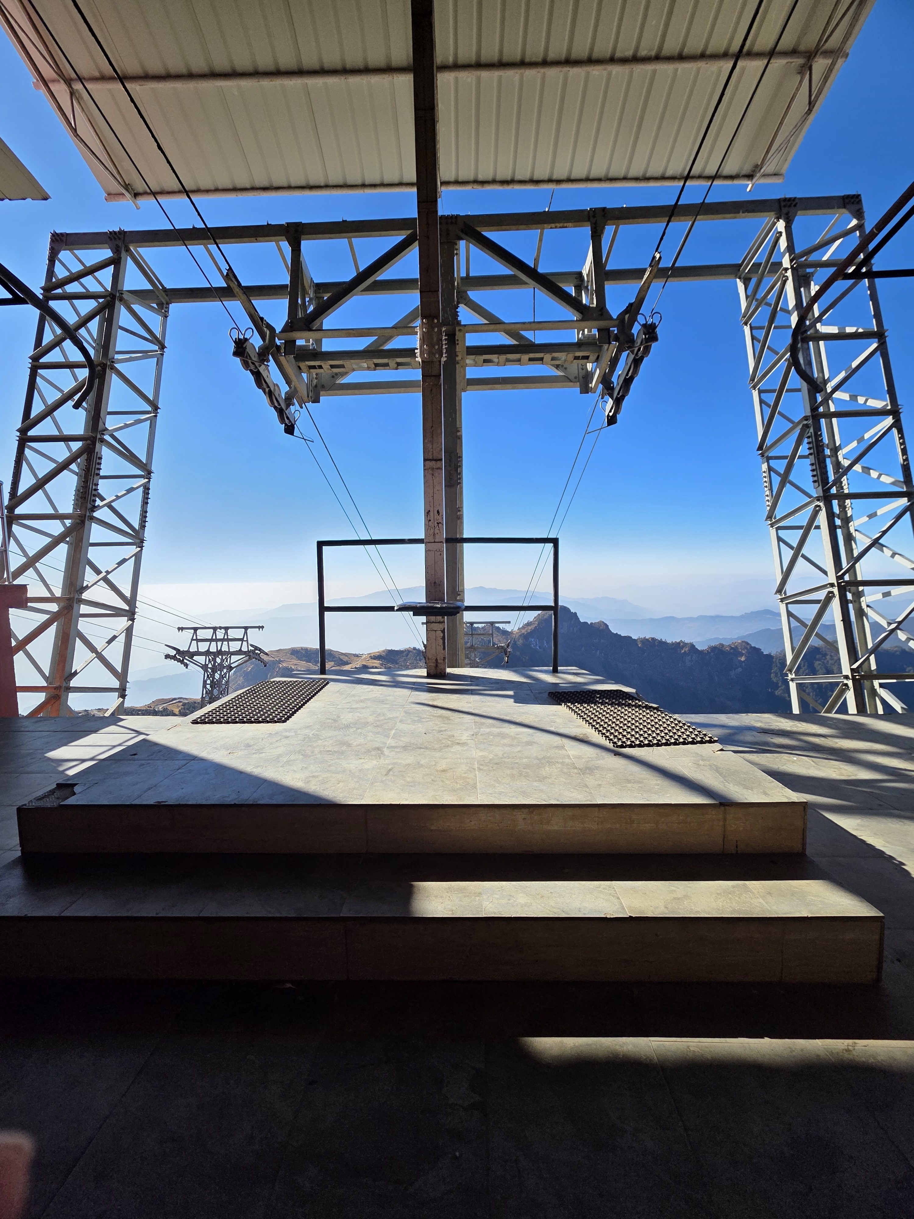 A zip line platform with metal framework overlooks distant mountains under a clear blue sky
