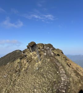 Stone steps trace a narrow mountain ridge toward rugged rock outcrops beneath a vast, vivid blue sky with wispy clouds and a faint daytime moon, creating a serene yet adventurous composition with distant hills on the horizon. 