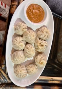 Ten steamed momos served on a white oval plate with a small bowl of Nepali-style tomato dipping sauce.
