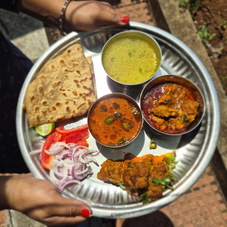 A traditional mutton thali served on a metal plate, featuring roti, flavorful meat curries, lentil soup, fresh tomato and onion slices, and a lime wedge. Held in hand in an outdoor setting, the meal showcases a rich and colorful spread of Indian cuisine.