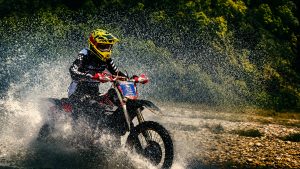 A motorcyclist in a yellow helmet speeds through a shallow water crossing, creating a dramatic spray. Lush green forest serves as the backdrop.
