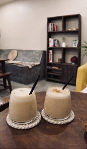 Two creamy iced coffees with black straws sit on coasters atop a wooden table. In the background, a cozy room with a bookshelf and seating area.