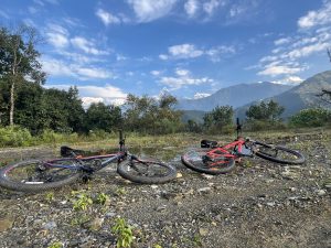 Two mountain bikes lie on a rocky path against a backdrop of lush trees and towering mountains under a vibrant blue sky with scattered clouds.
