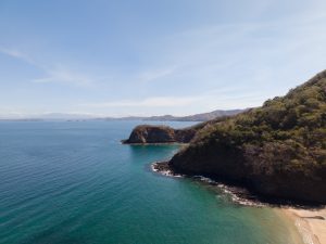 An aerial view of a turquoise bay bordered by rocky coastal cliffs and tree-covered hills. A small sandy beach appears at the edge of the water, with distant islands and mountains visible under a clear blue sky.