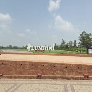 Brick platform with large "Lumbini Nepal" sign set against a backdrop of lush greenery and a partly cloudy sky, conveying a serene.