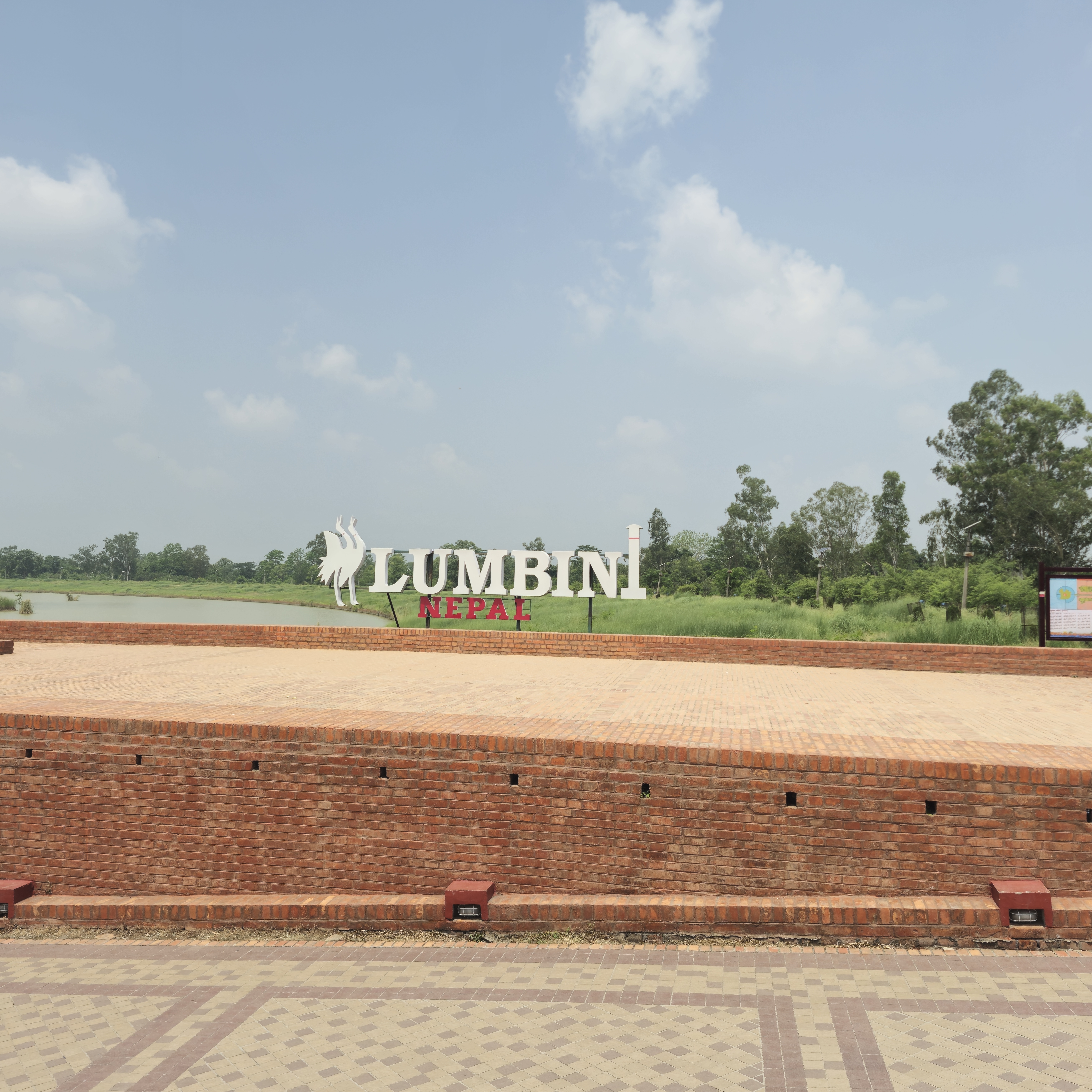 Brick platform with large "Lumbini Nepal" sign set against a backdrop of lush greenery and a partly cloudy sky, conveying a serene.
