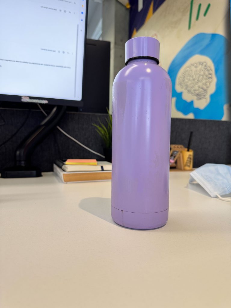 A purple water bottle on a white desk, near a computer monitor, notebooks, a small plant, and a face mask.