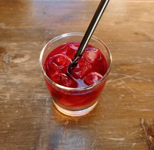 A clear glass filled with a translucent, vibrant red beverage, several square ice cubes, and a single thick black plastic straw. 
