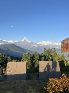 Two tents are set up in a scenic campsite with the snow-capped Himalayas in the background. A wooden post with a heart is visible, evoking a sense of adventure and tranquility.