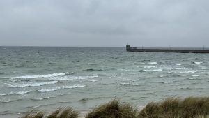 A stormy coastline with choppy waves and a foamy shoreline stretches beneath a heavy gray sky. A graffiti-lined pier with small lights extends into the sea, while light brown grasses sway in the foreground wind.
