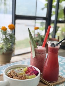 A colorful smoothie bowl topped with walnuts, almonds, kiwi, and pomegranate is next to a pink smoothie and red juice, set on a table with a scenic view.