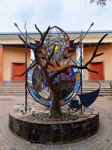 A massive circular stained-glass installation built with red, blue, yellow, and checkered mosaic panels, flanked by two black metal spears and set behind a mature tree.