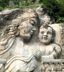 A large stone relief sculpture carved into a rock face shows an adult with flowing hair gazing affectionately at a child with expressive features, framed by a carved balustrade. Greenery surrounds the natural rock setting. 