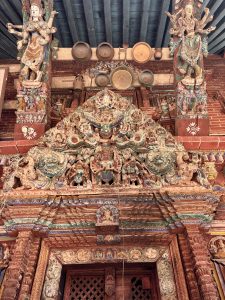 An intricately carved and painted wooden tympanum (torana) above a temple doorway depicting Bhairava and multiple deities, flanked by painted deity columns with brass ritual vessels displayed above, Nepal. 