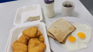 A hearty breakfast featuring fried eggs and toast on a white plate, hash browns in a takeout box, a cup of tea, and a bottle of yogurt drink.