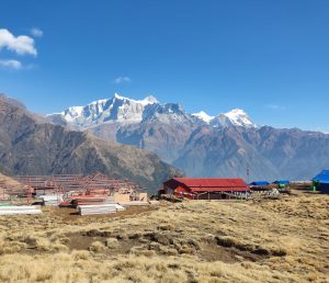 Mountain landscape with snow-capped peaks under a clear blue sky. Foreground features a red-roofed building, colorful flags, and dry grass. Calm and majestic.
