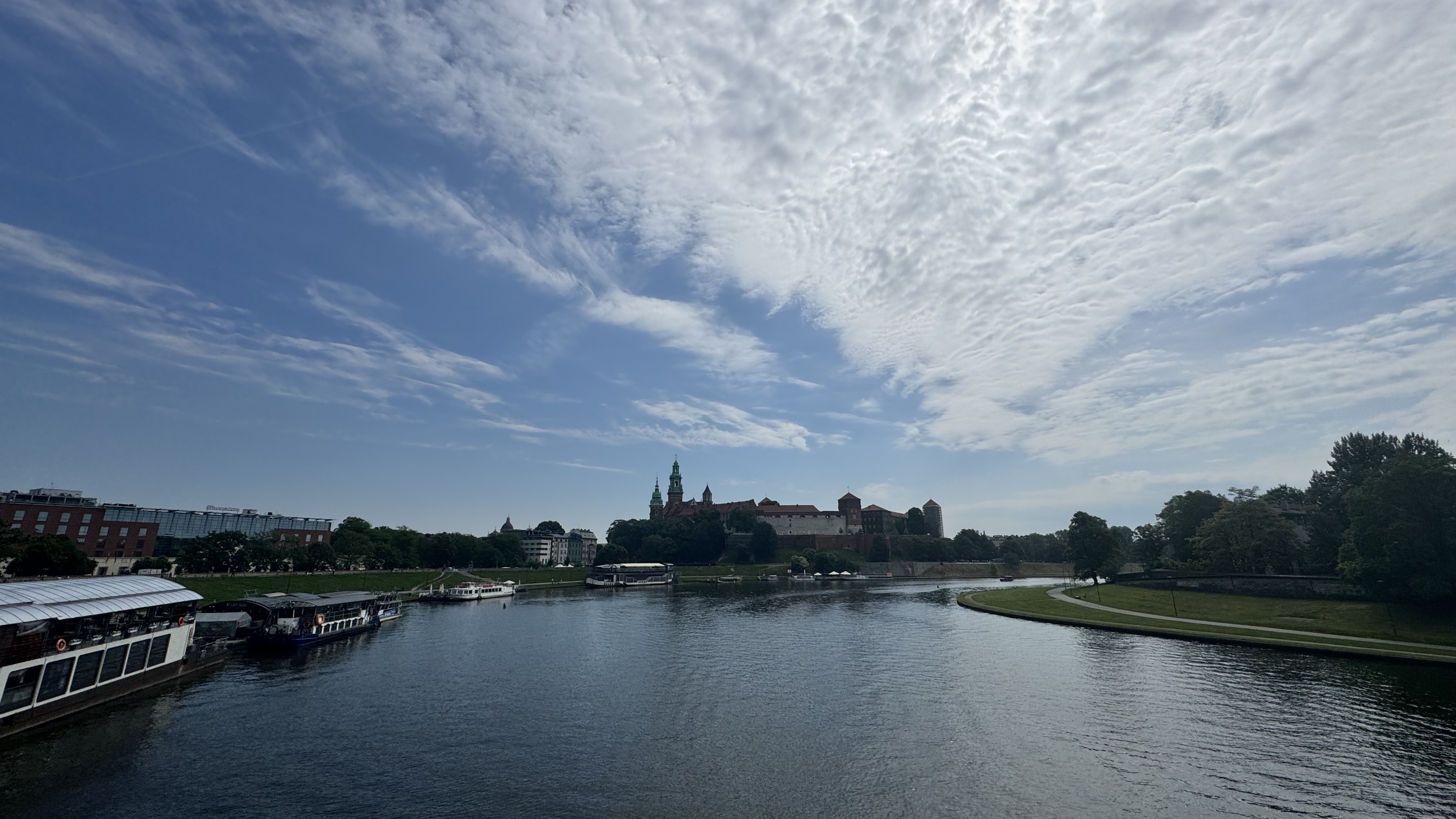 A calm river under a blue sky with light clouds. Boats are docked on the left bank, including one with a white roof. On the right, there is a grassy area with trees, and across the river stands a castle with towers and green spires.