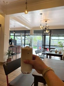 A hand holding a tall glass of iced coffee with a clear straw inside a bright, modern café. Wooden tables, dark chairs, and stylish hanging lights fill the cozy background. 