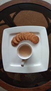 A white square plate holds a cup of coffee and a crescent of round biscuits.