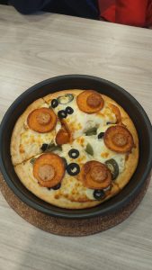 A freshly baked pan pizza  with melted cheese, olives, sliced sausages, and bell peppers is in a black baking pan on a light wooden table.
